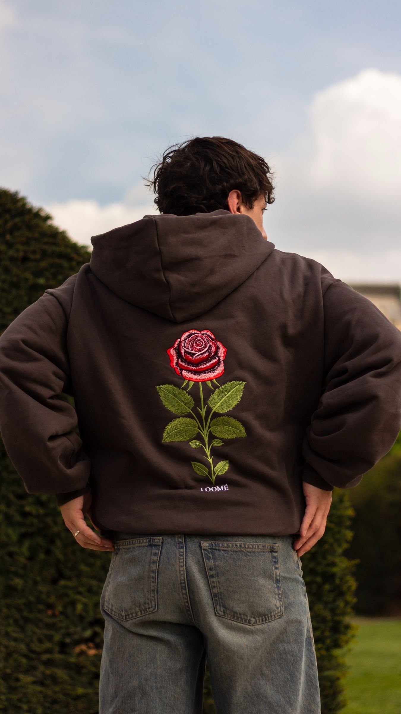 Ein Mann von hinten fotografiert im Ash-Grey-Hoodie, die Hände in die Hüften gestemmt, die gestickte Rose füllt die Mitte seines Rückens. Links von ihm steht eine geschnittene Hecke, dahinter Gras und blasse Wolken.