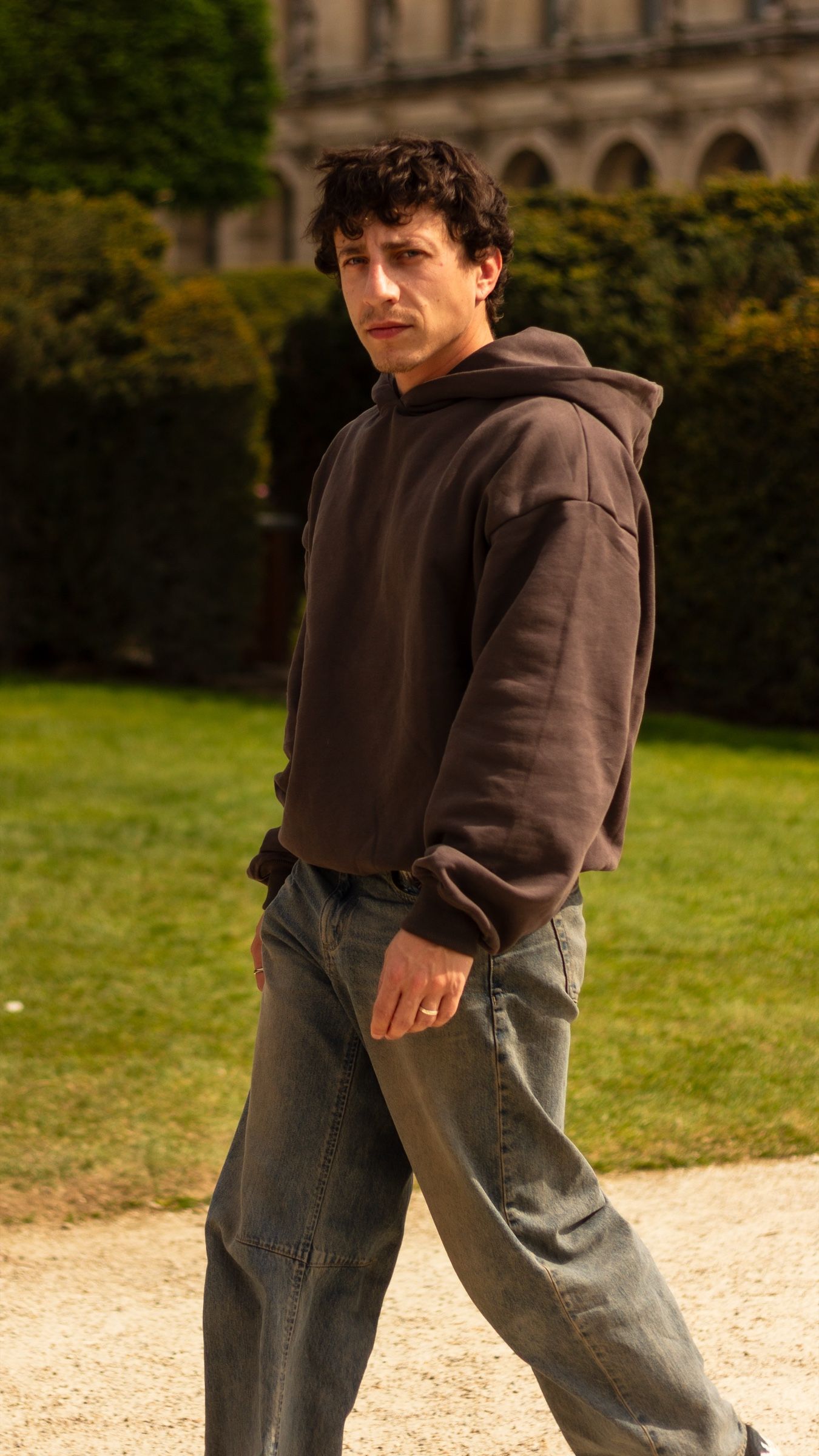 Le même homme marchant sur une allée de gravier en hoodie Ash Grey et jean large délavé, tourné vers l'appareil. Des haies taillées et une façade parisienne en pierre pâle sont derrière lui, au soleil.