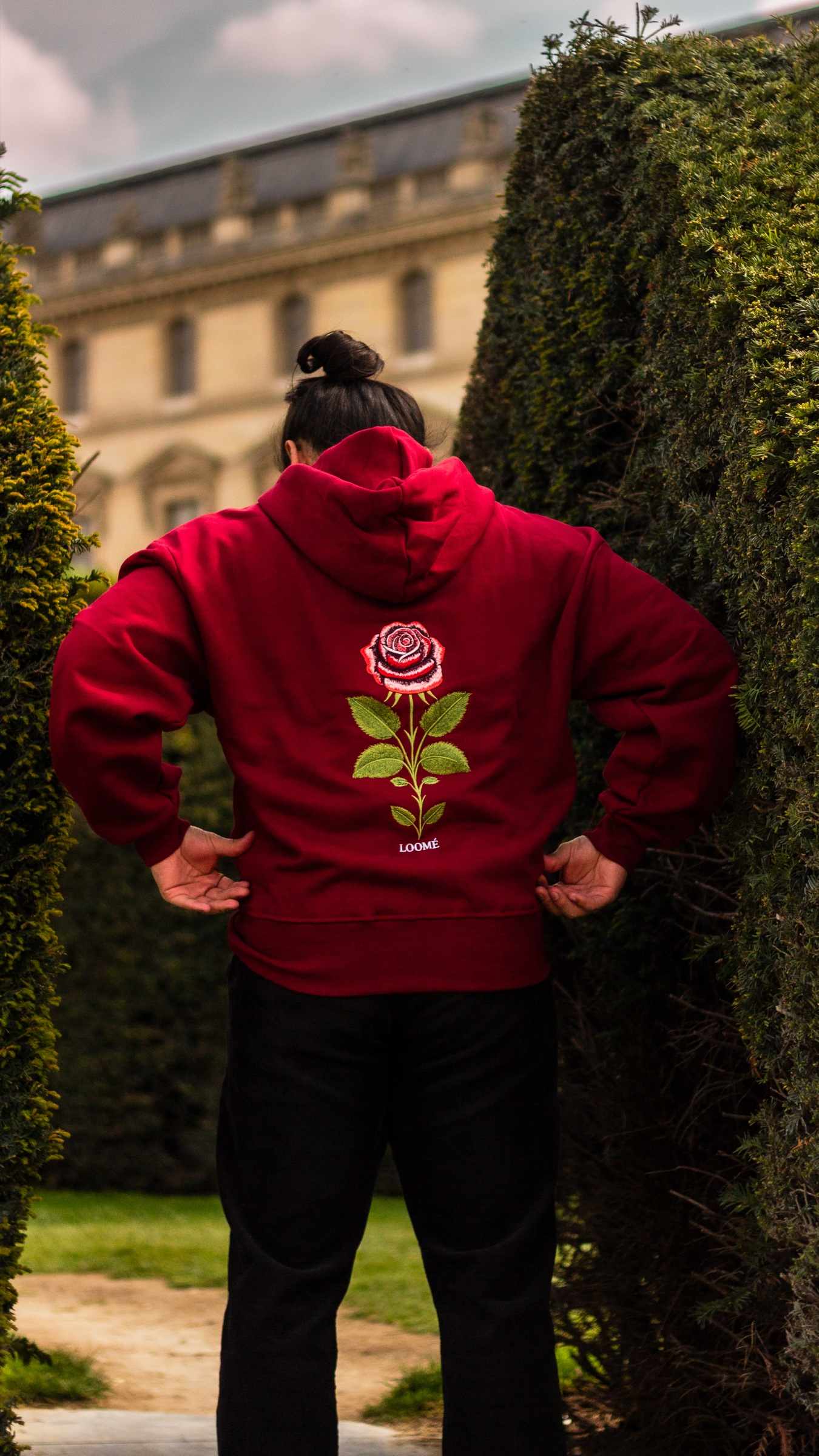Une personne vue de dos en hoodie Burgundy, tête baissée, debout entre deux hautes haies taillées. La rose descend au centre du dos, et une façade de palais en pierre pâle s'élève au loin.