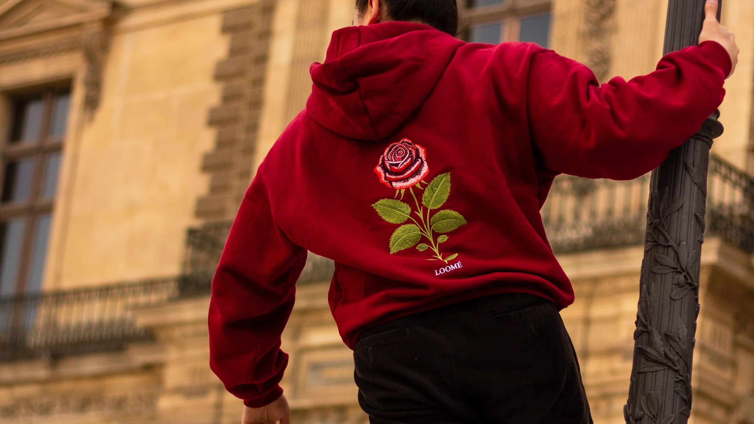 Ein enger Ausschnitt des Burgundy-Hoodies von hinten, ein Arm ist erhoben und hält eine Laterne, der Ballonärmel fällt in Falten. Die Rose und die LOOMÉ-Stickerei sind scharf, der Steinbalkon dahinter unscharf.