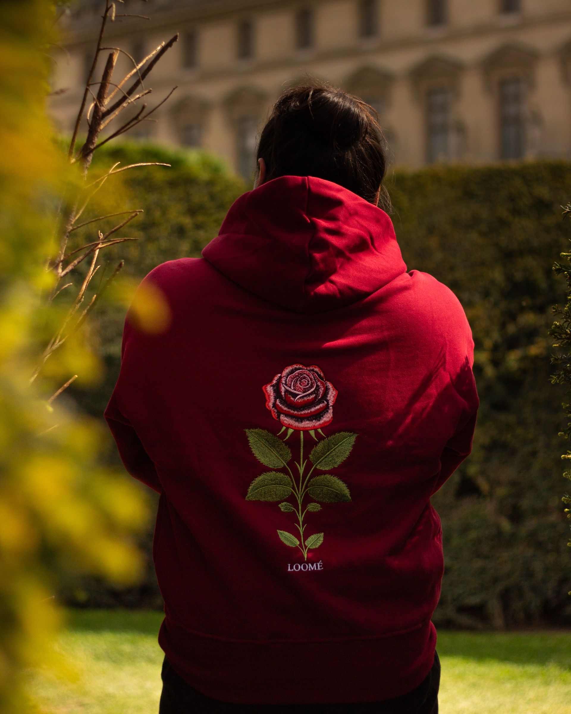 Une personne de dos en hoodie Burgundy devant une haie taillée, avec des feuilles jaunes floues traversant le premier plan. La rose et sa longue tige descendent au centre du dos.