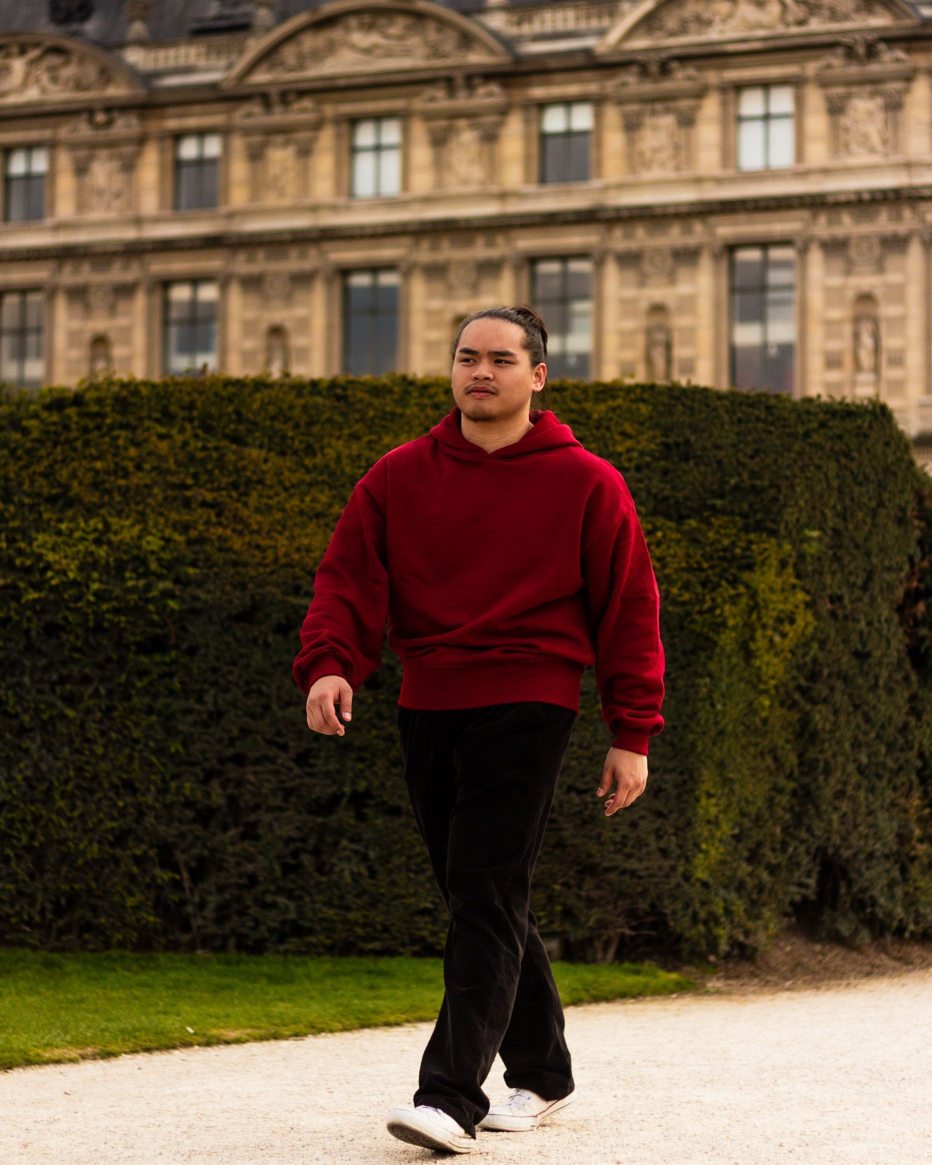 Ein Mann geht auf einem Kiesweg auf die Kamera zu, im Burgundy-Hoodie, schwarzer Hose und weißen Sneakern. Die Vorderseite des Hoodies ist leer, das Bündchen am Saum sitzt an der Hüfte, im Hintergrund füllen eine Hecke und eine Palastfassade das Bild.