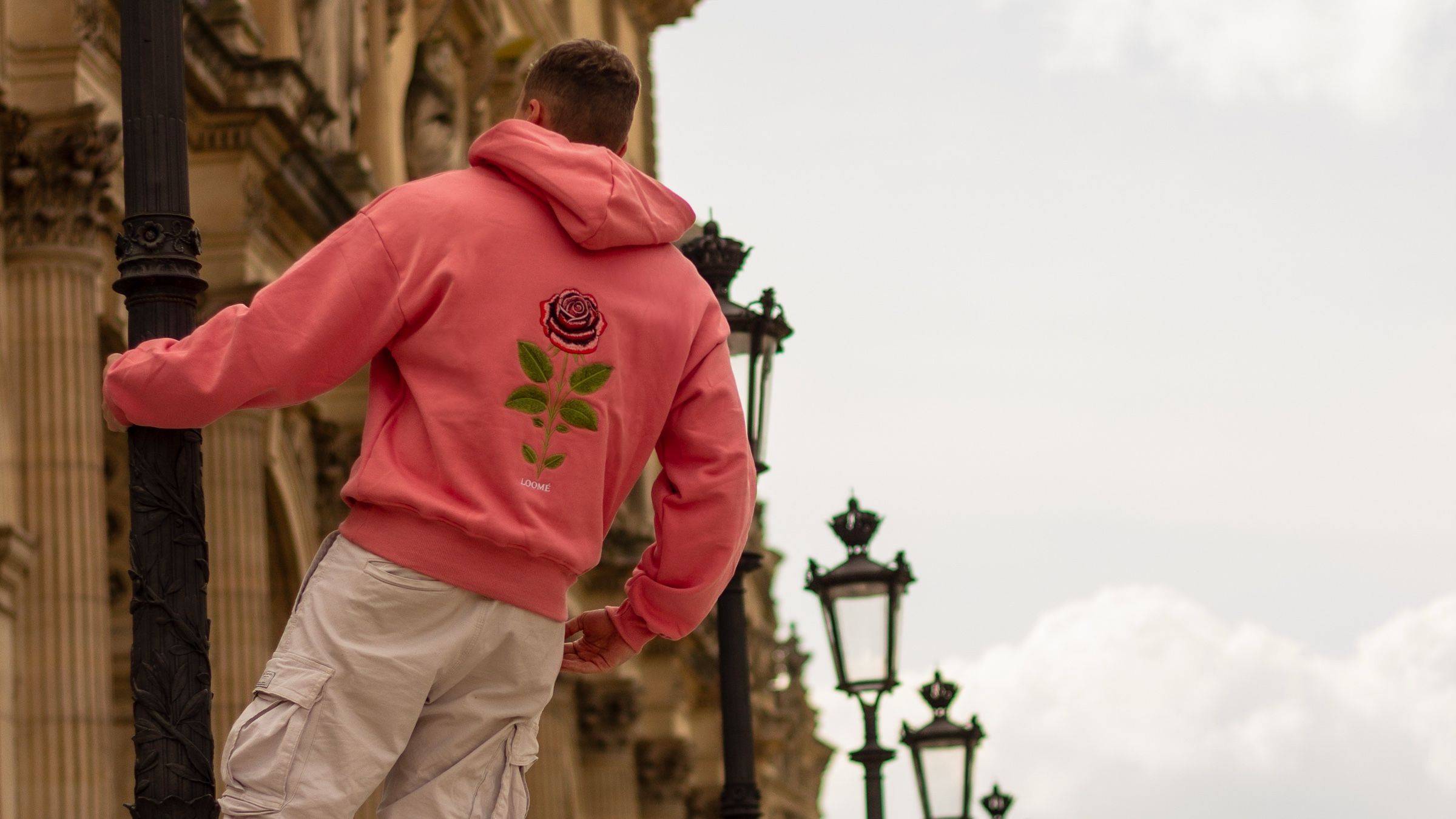 A man leaning on a lamppost with his back to the camera, in the Bubblegum Pink hoodie and off-white cargo trousers. The rose is centred on his back, with Paris lampposts and pale stone behind.