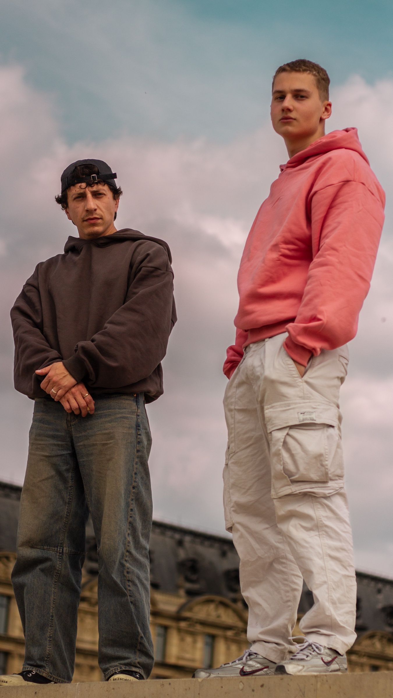Deux hommes debout sur un rebord, photographiés en contre-plongée devant un ciel nuageux. À gauche le hoodie Ash Grey, bras croisés et jean large ; à droite le hoodie Bubblegum Pink, mains dans les poches.