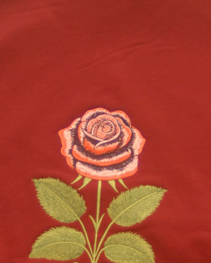 Eine rote Rose, auf bordeauxfarbenen Stoff gestickt, aus der Nähe: die Blüte in Rottönen und Rosa gearbeitet, darunter olivgrüne Blätter an einem langen Stiel.