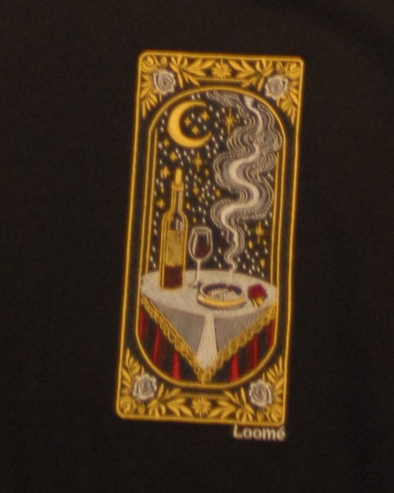 Ein gesticktes Feld auf einem schwarzen Hoodie: ein goldener Rahmen um eine Nachtszene mit Mondsichel, Sternen, einer Weinflasche, einem gefüllten Glas und einem gedeckten Tisch, darunter die Signatur Loomé.